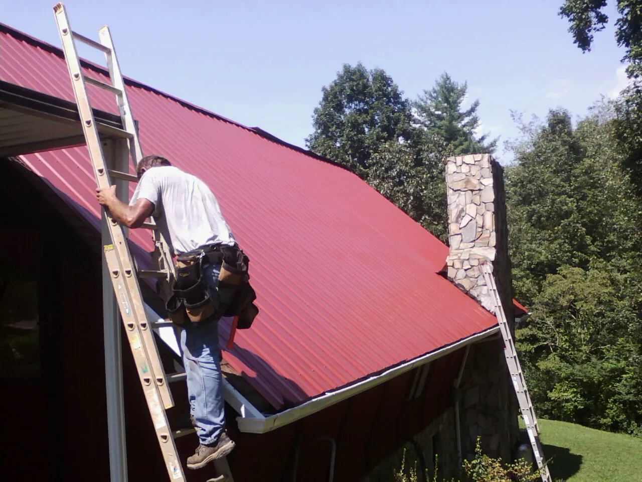 Roofer on a ladder beside a red metal roof and stone chimney — Soffit & Fascia Repair by Roofing Brothers in Giddings